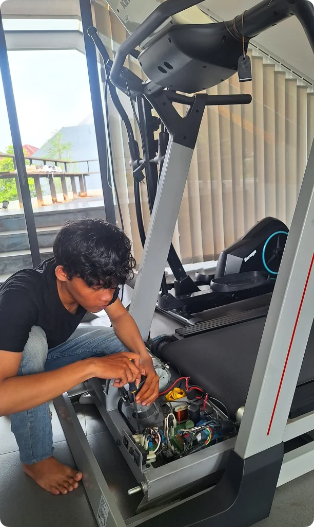 Teknisi muda sedang melakukan perbaikan kelistrikan pada mesin treadmill di lokasi pelanggan